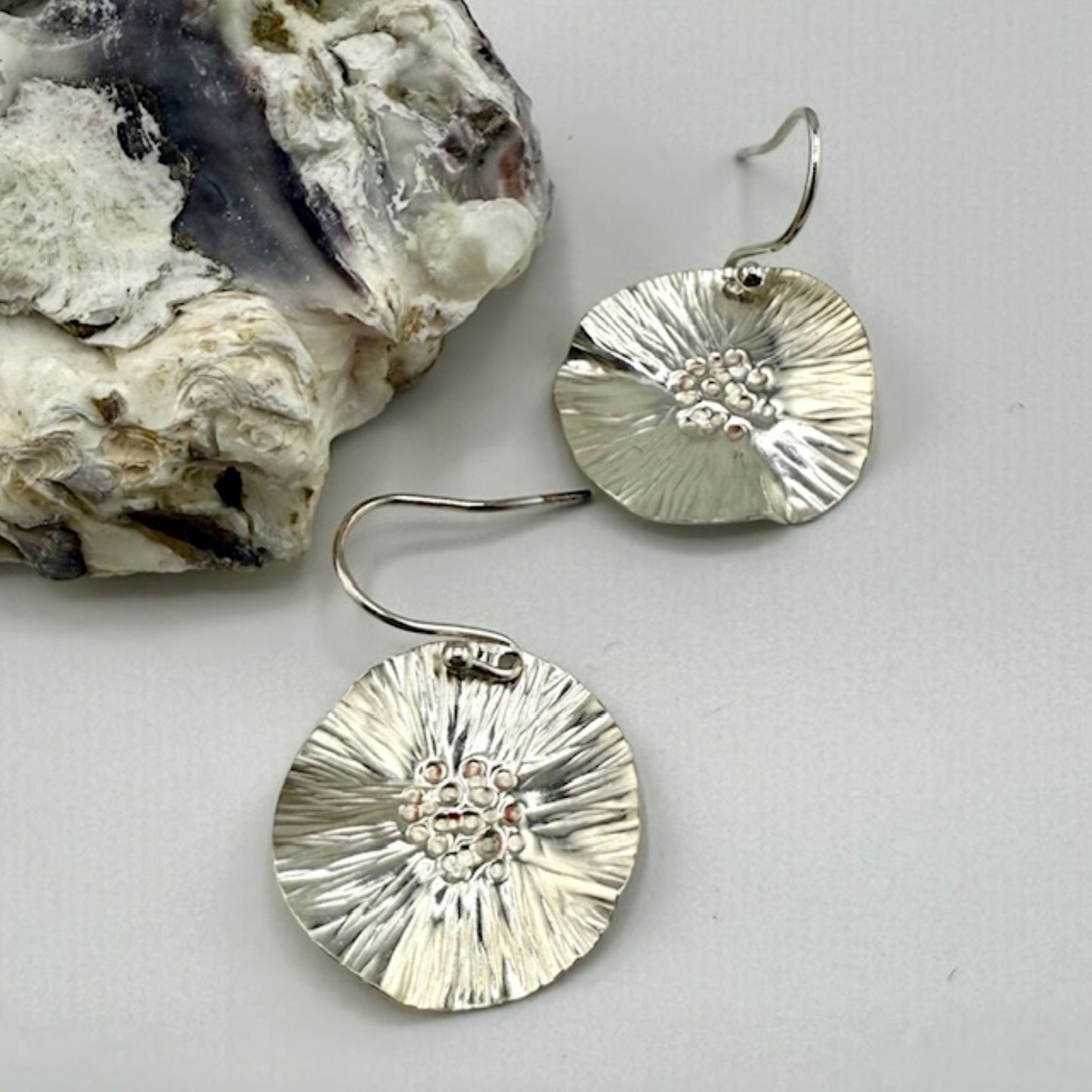 Silver textured earrings on a light gray background with a piece of rock.