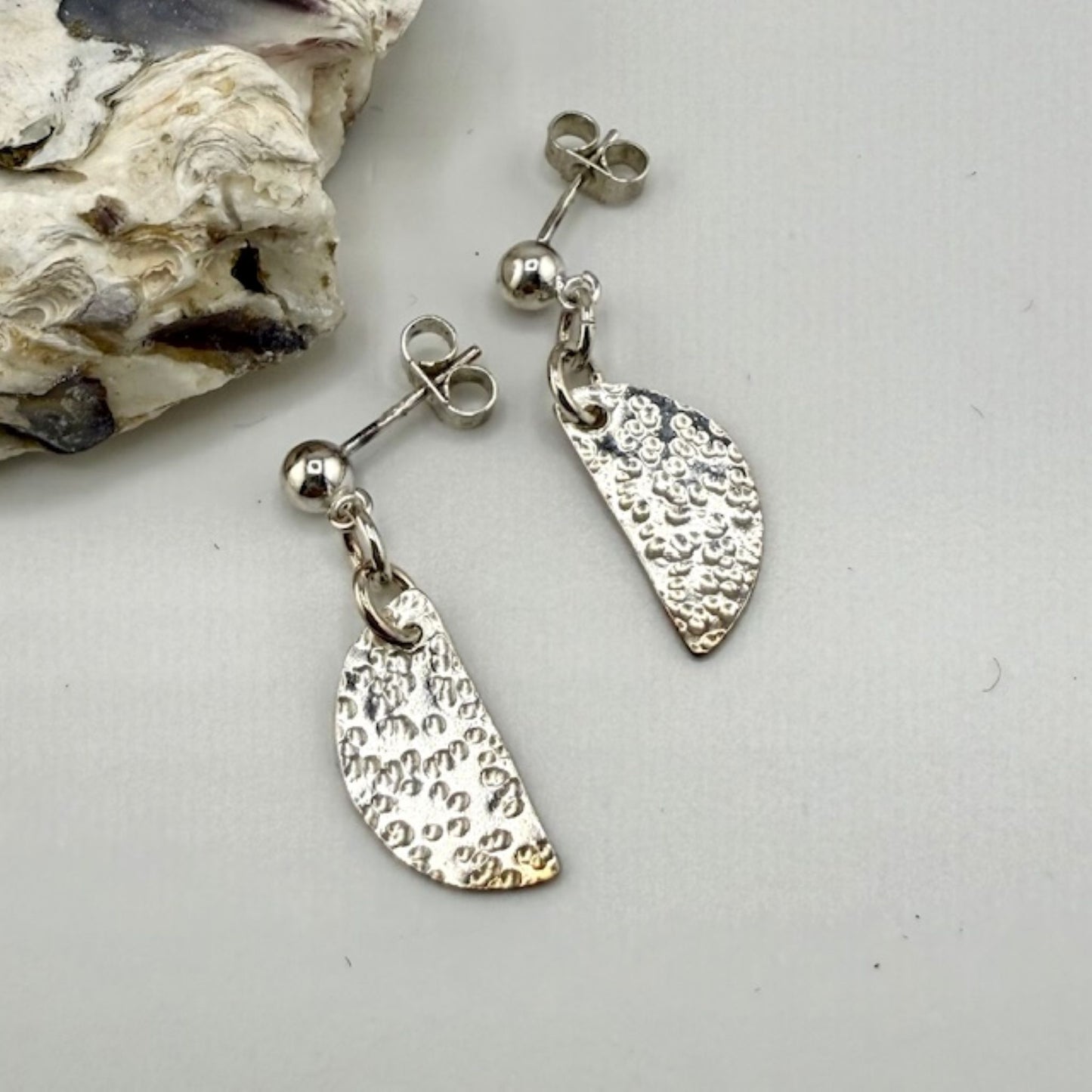 Silver half moon stud earrings on a light gray background with a shell.