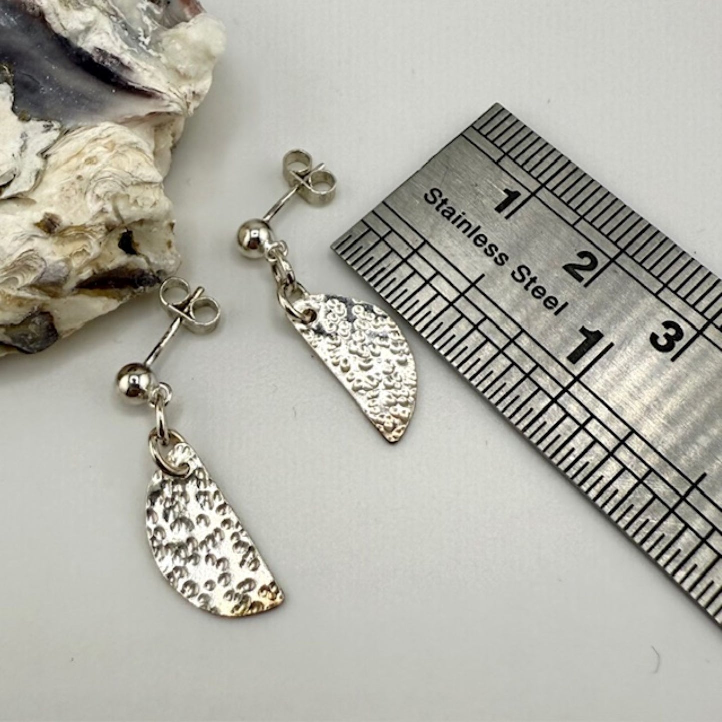 Silver half moon earrings with a textured design next to a ruler on a white surface