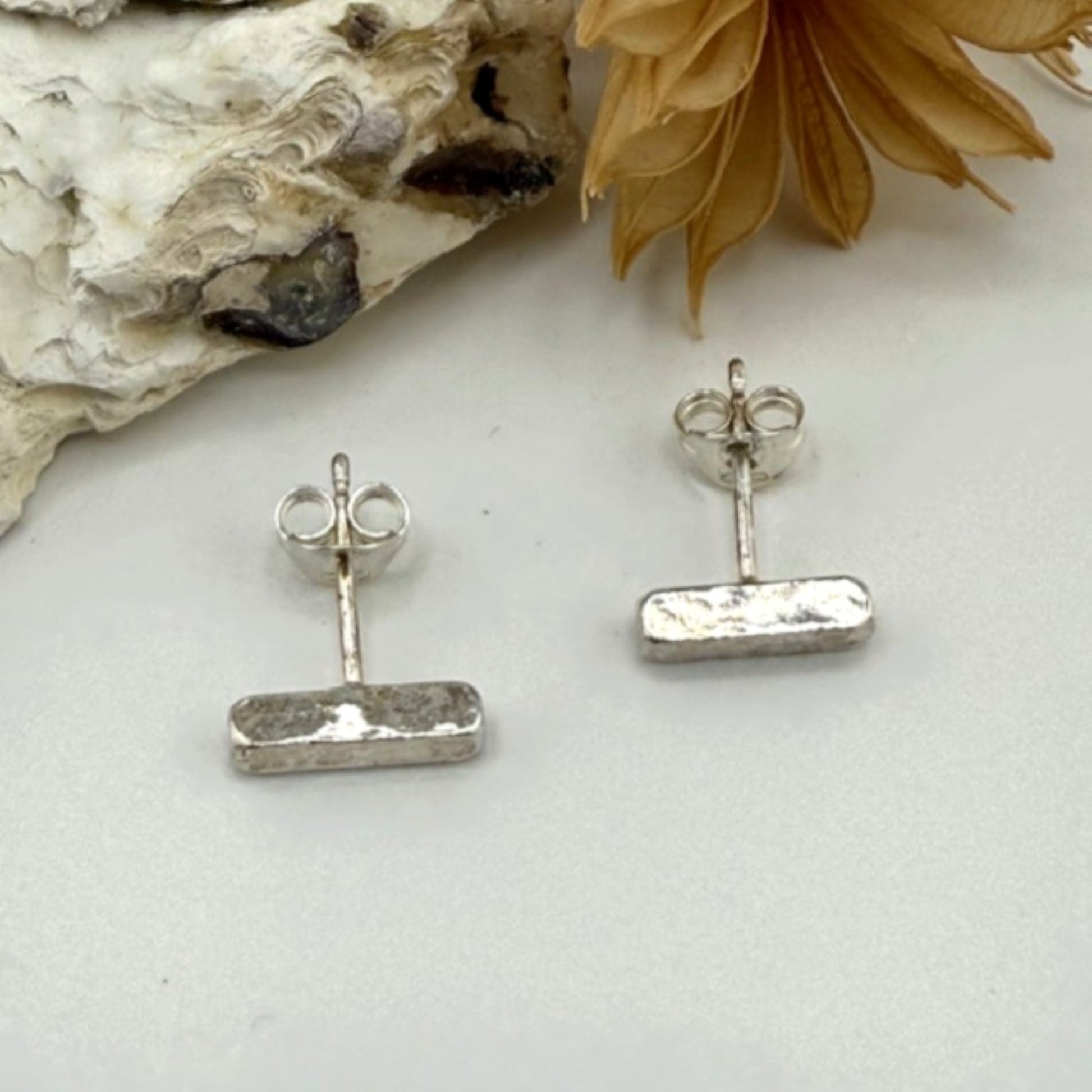 Silver bar stud earrings on a white surface with a natural rock and flower in the background