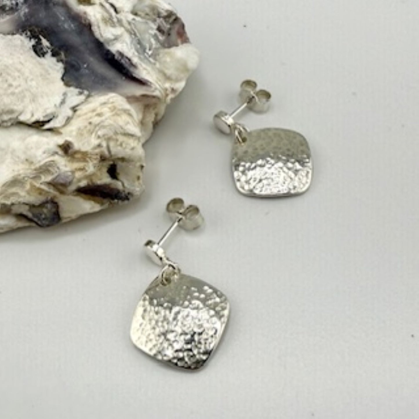 Pair of silver square earrings on a white surface with a rock in the background