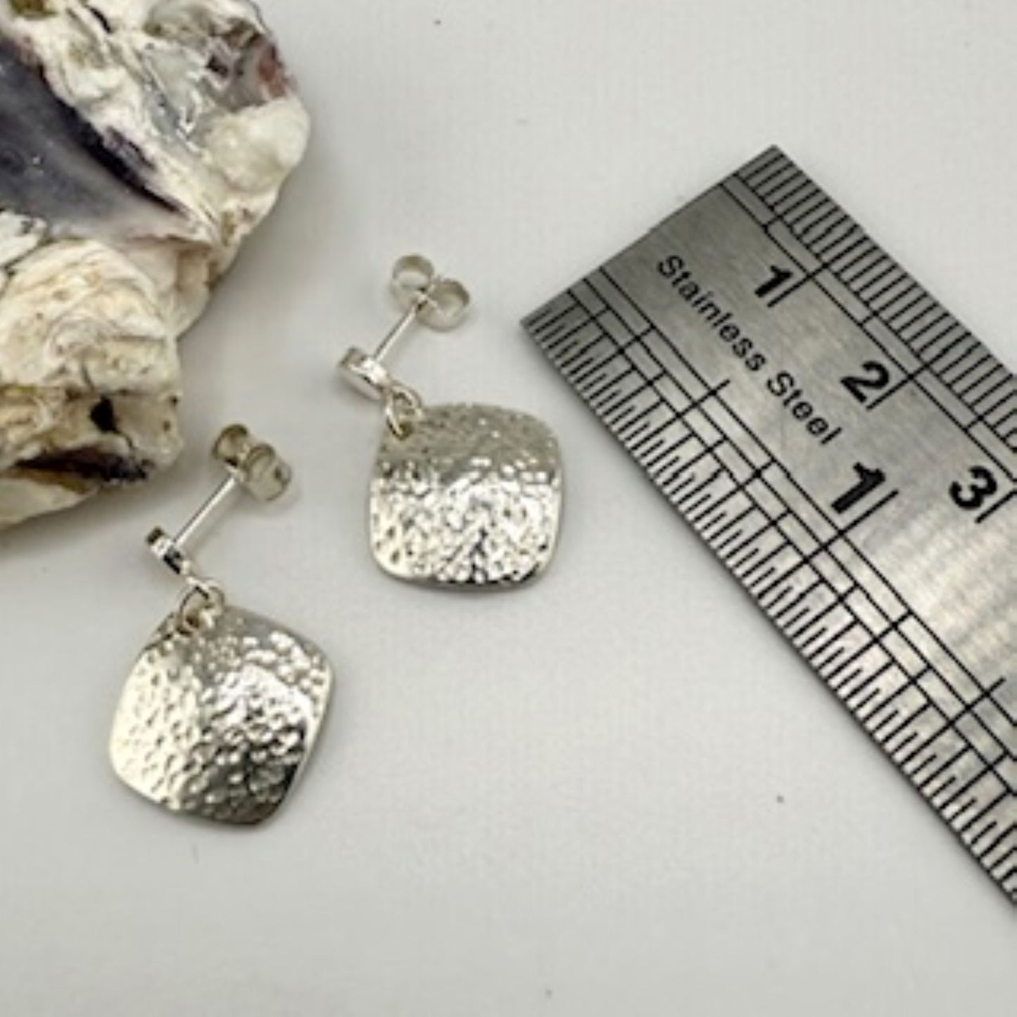 Silver square earrings with textured design on a white surface with a ruler for scale.