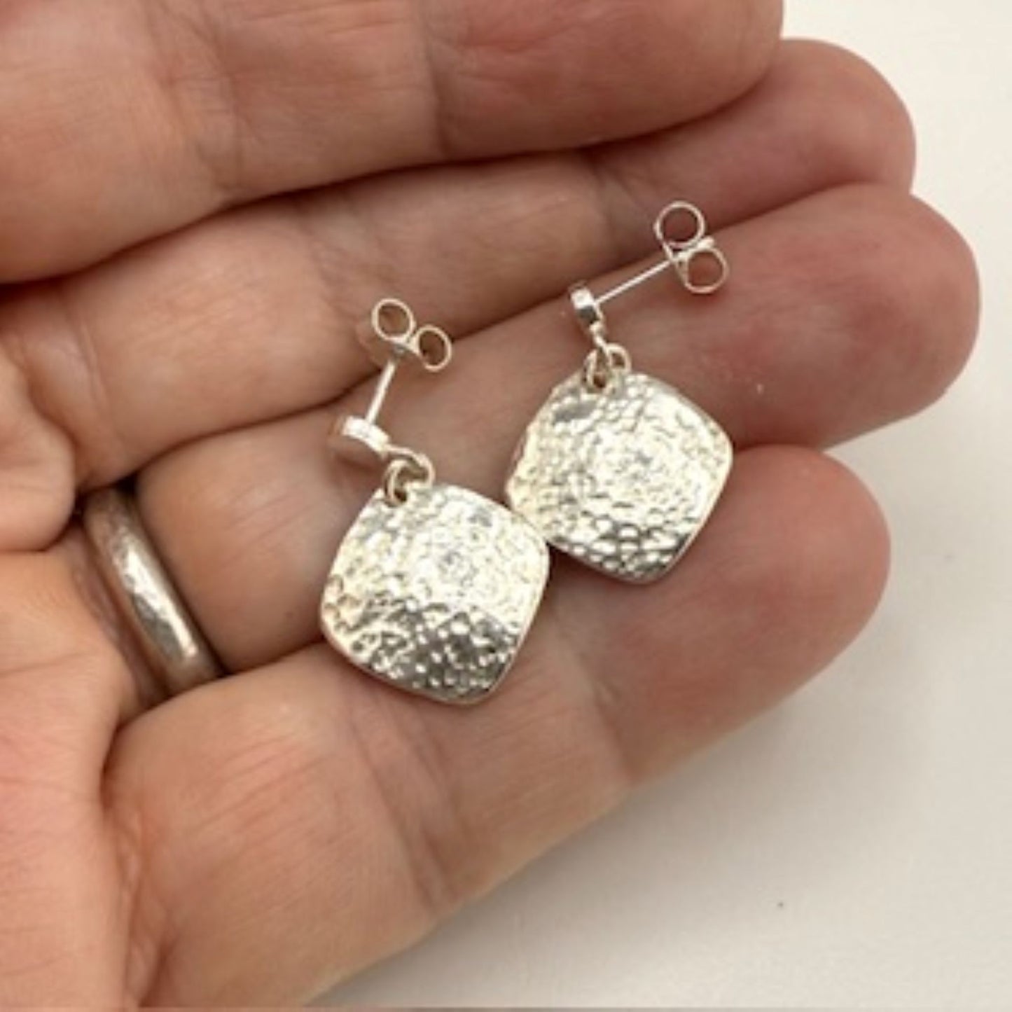 Silver square earrings held in a hand against a white background