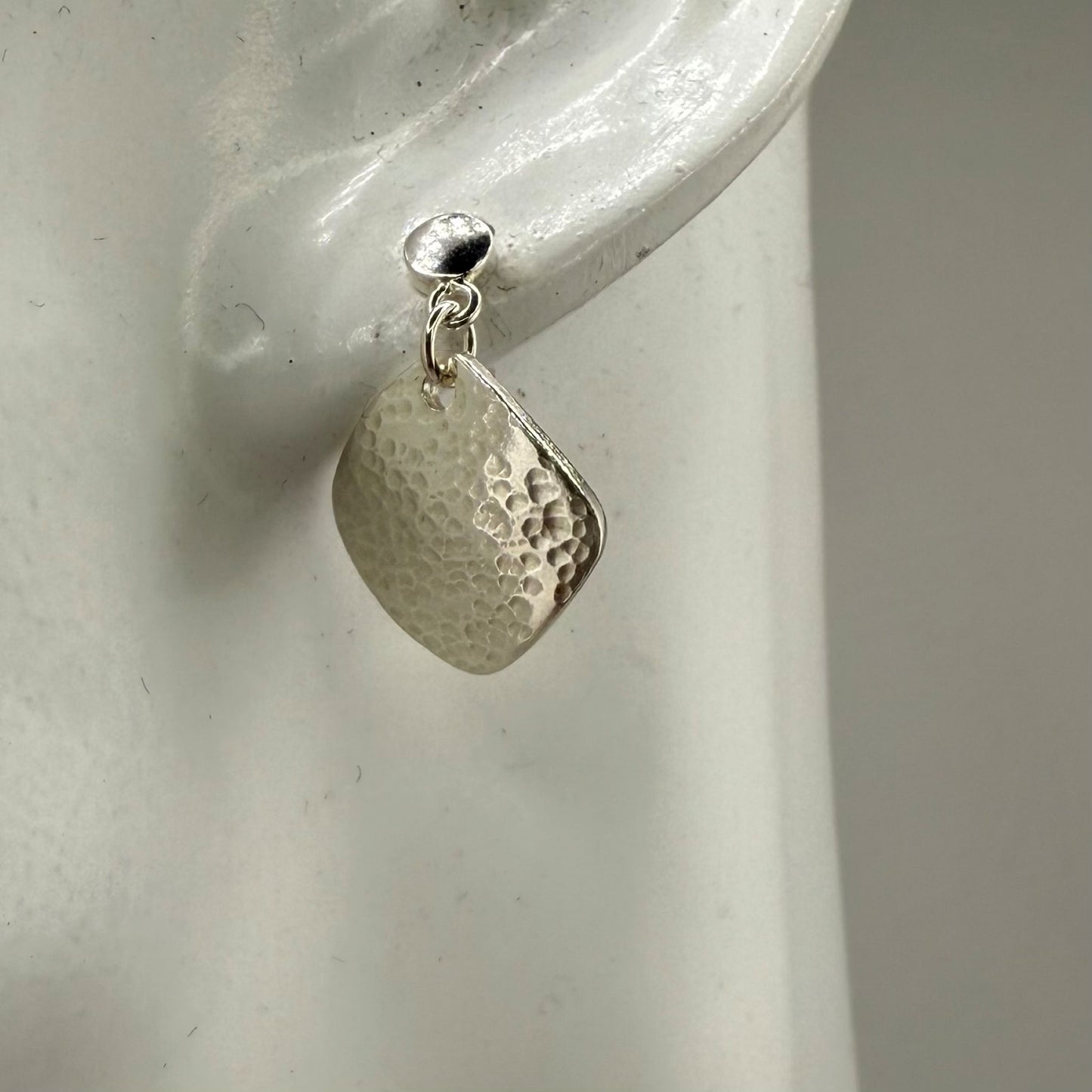 Silver square earring with textured teardrop design on a white ear display
