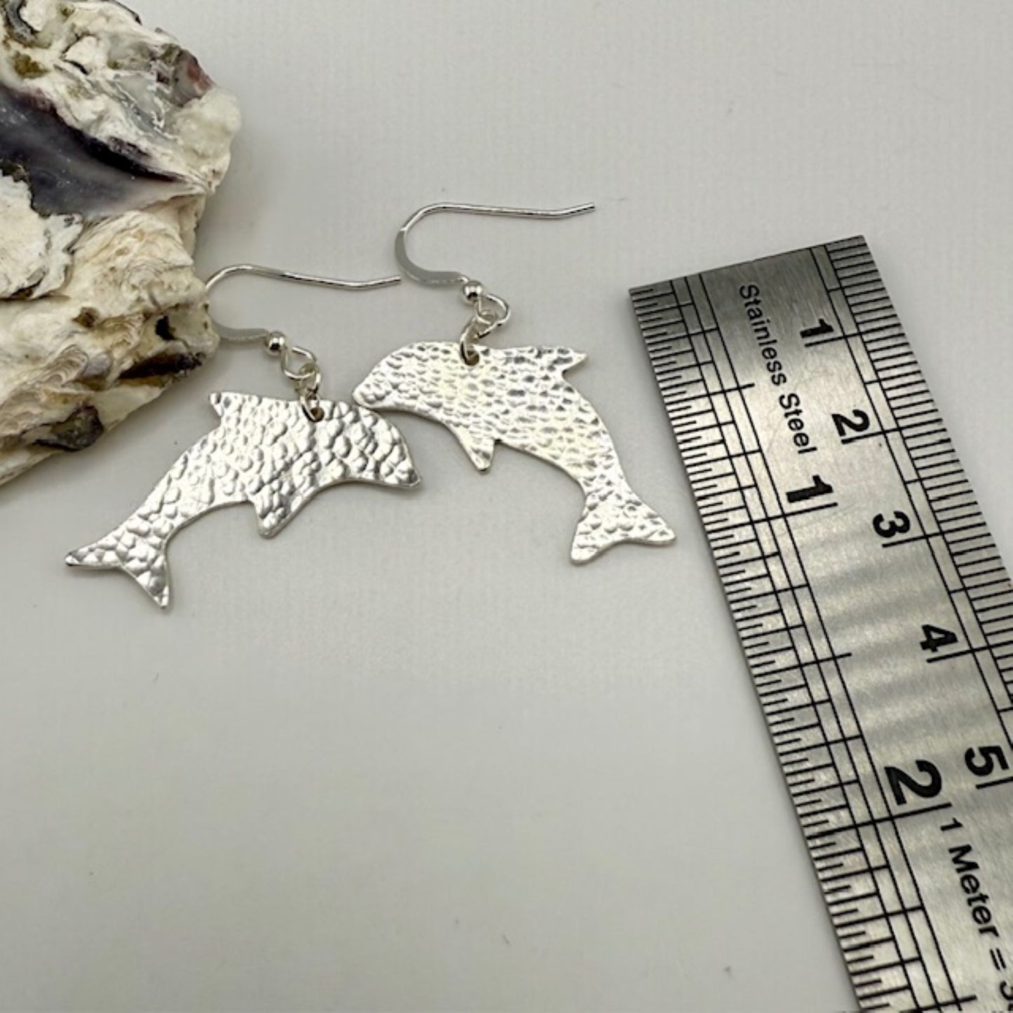 Silver dolphin-shaped earrings with a ruler for scale on a white background