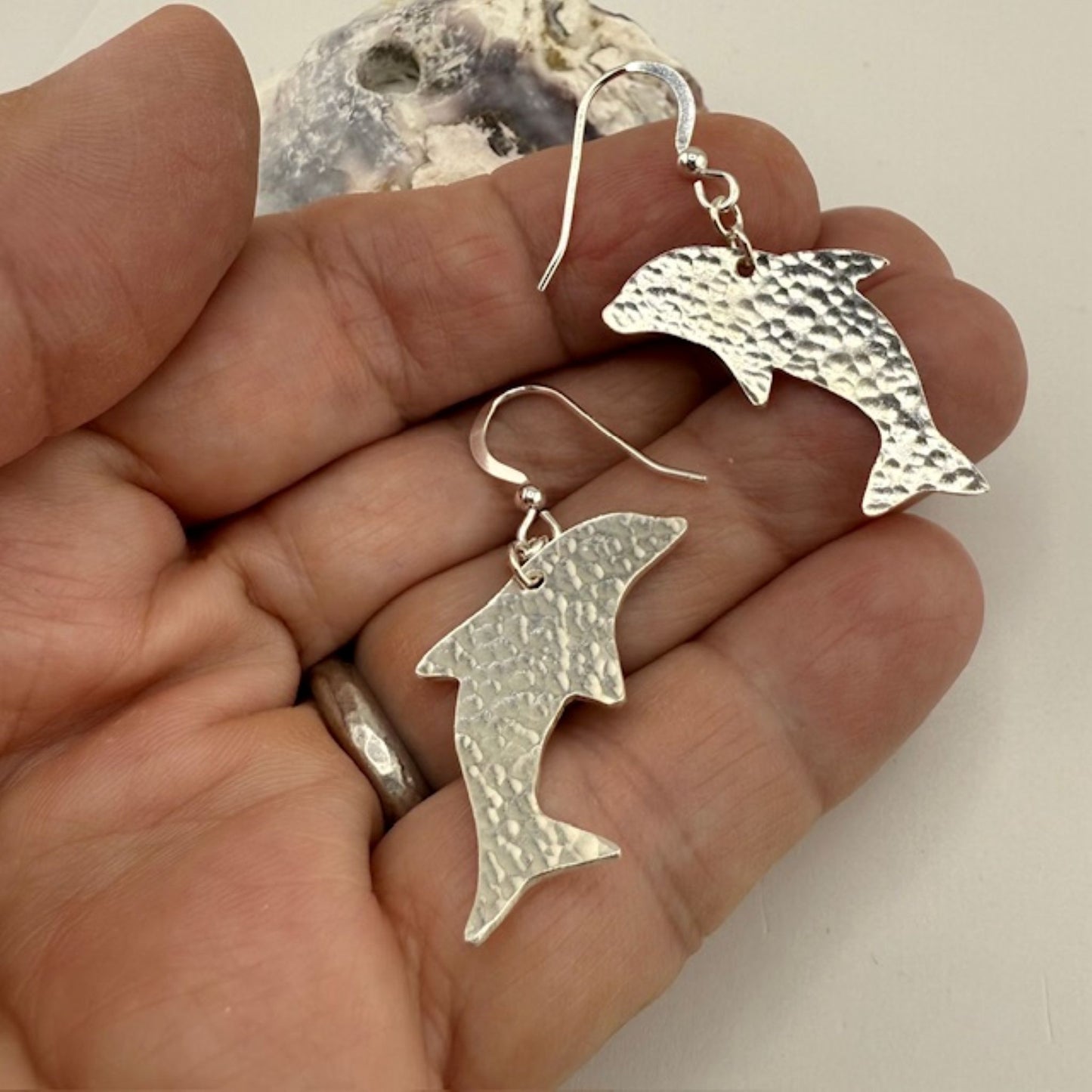 Silver dolphin earrings held in a hand against a neutral background