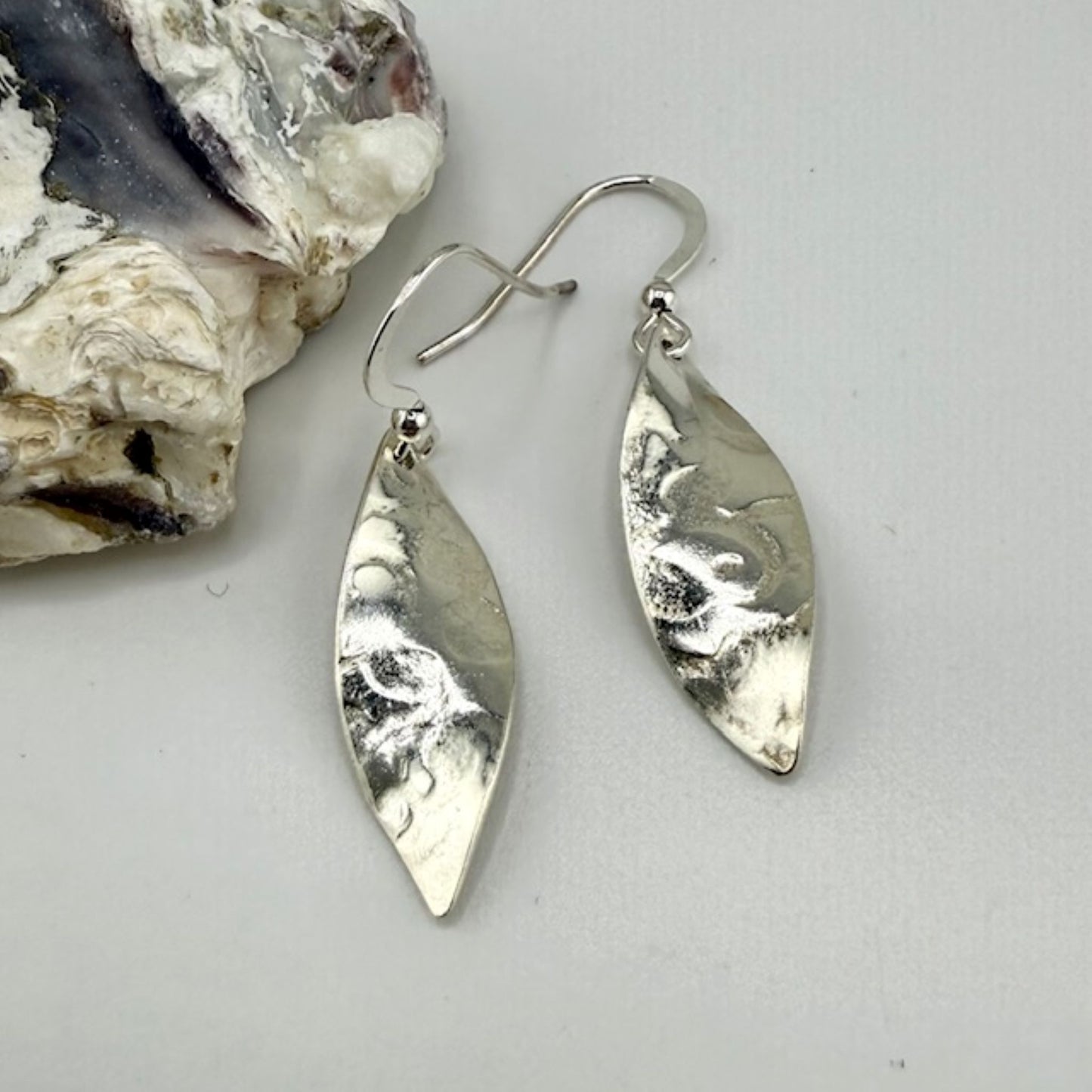 Silver leaf earrings on a light gray background with a shell.