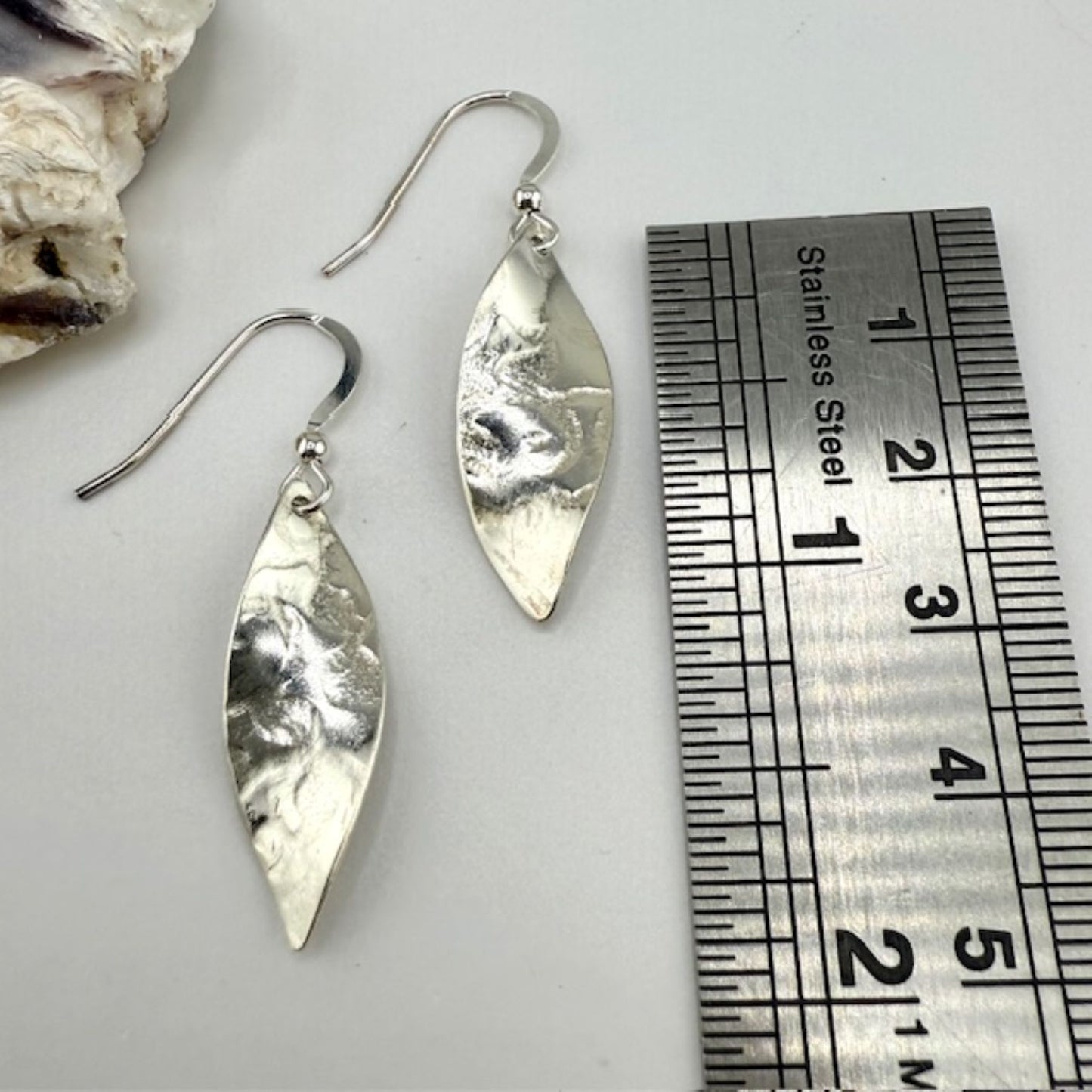 Silver leaf-shaped earrings next to a ruler on a light gray background