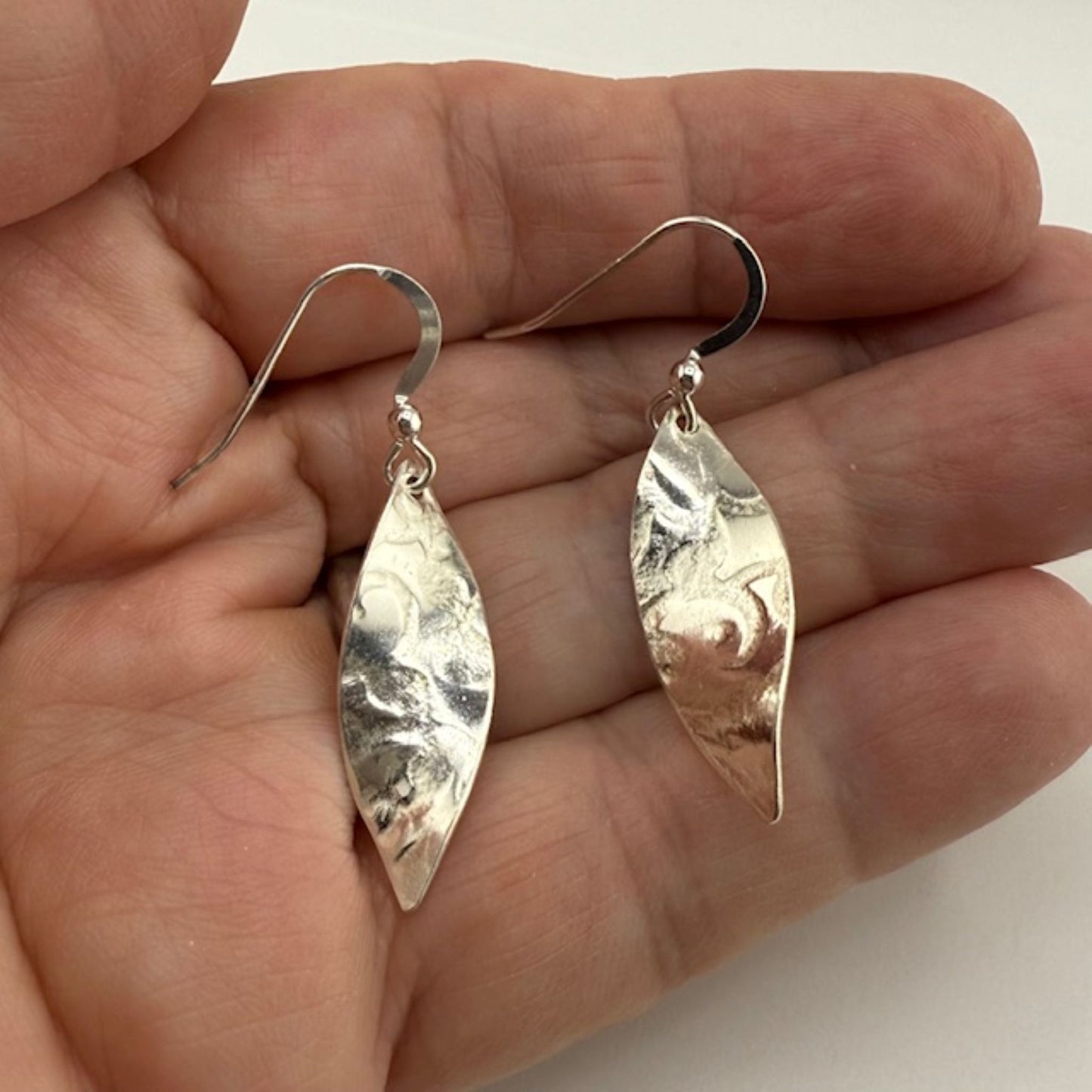 Silver leaf-shaped earrings held in a hand against a neutral background