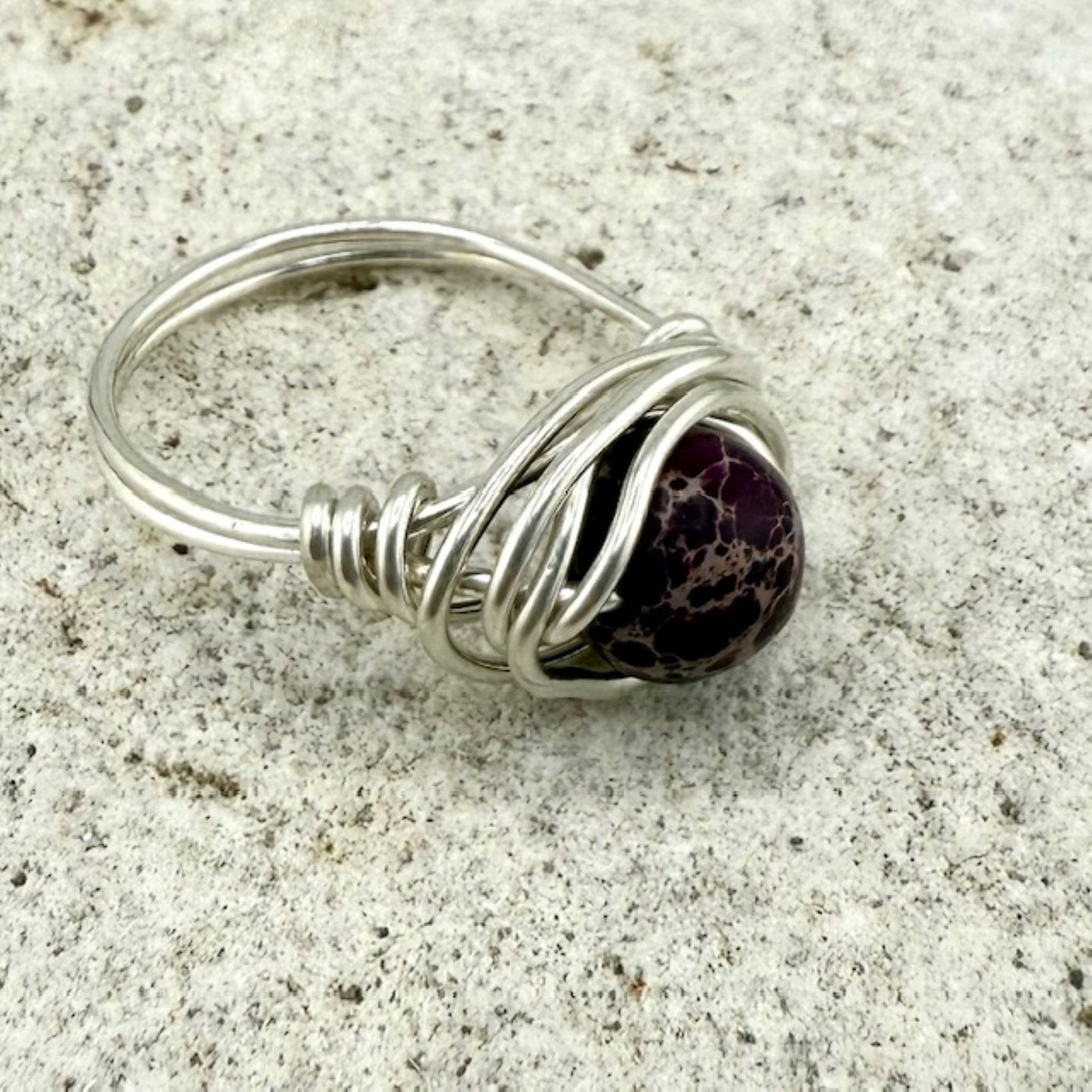 Silver wire-wrapped ring with a purple jasper gemstone on a textured surface