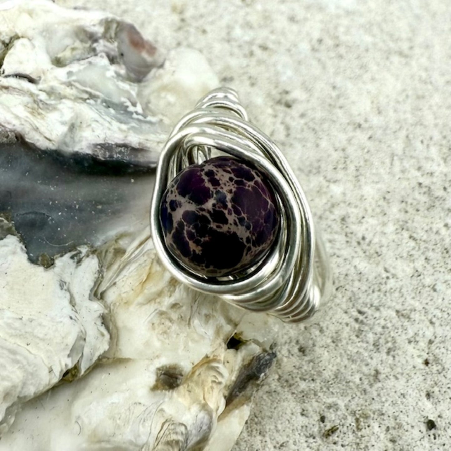 Silver wire-wrapped ring with a purple speckled jasper gemstone on a textured surface
