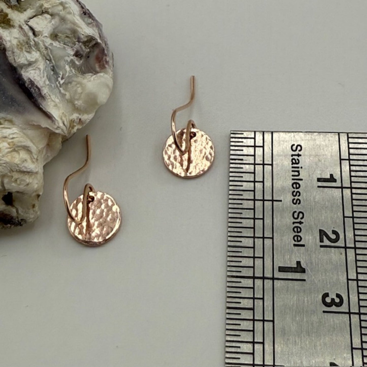 Rose gold earrings next to a ruler for size reference on a neutral background