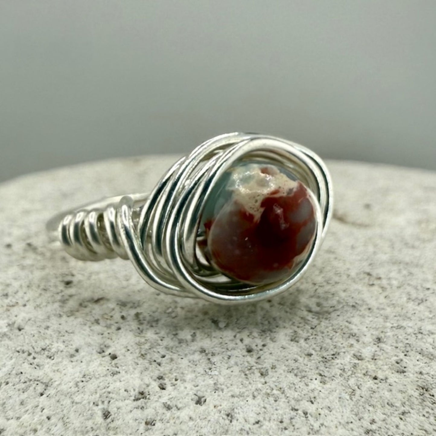 Silver wire-wrapped ring with a sea sediment jasper stone on a white stone surface