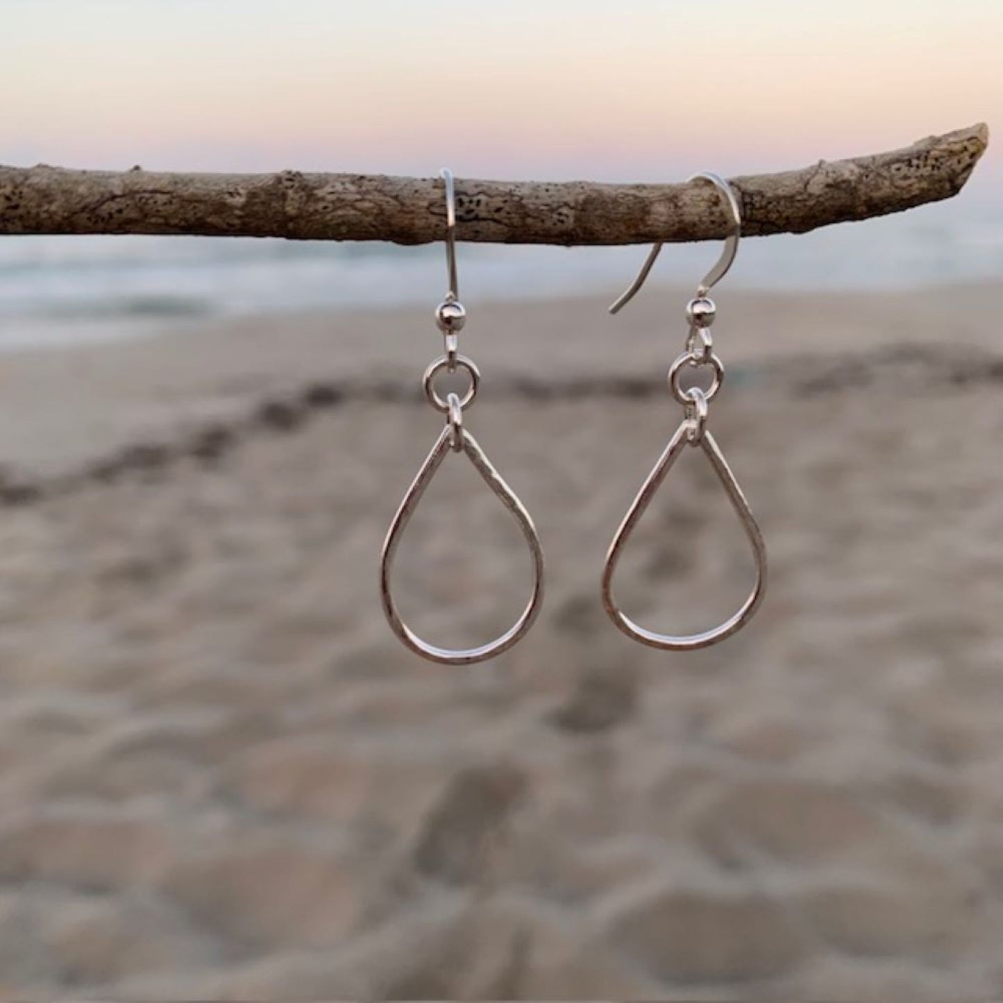 Teardrop-shaped sterling silver earrings hanging on a branch with a beach background