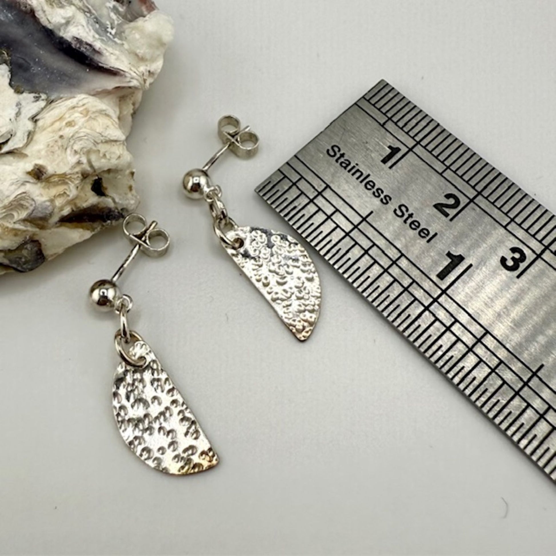Silver half moon earrings with a textured design next to a ruler on a white surface