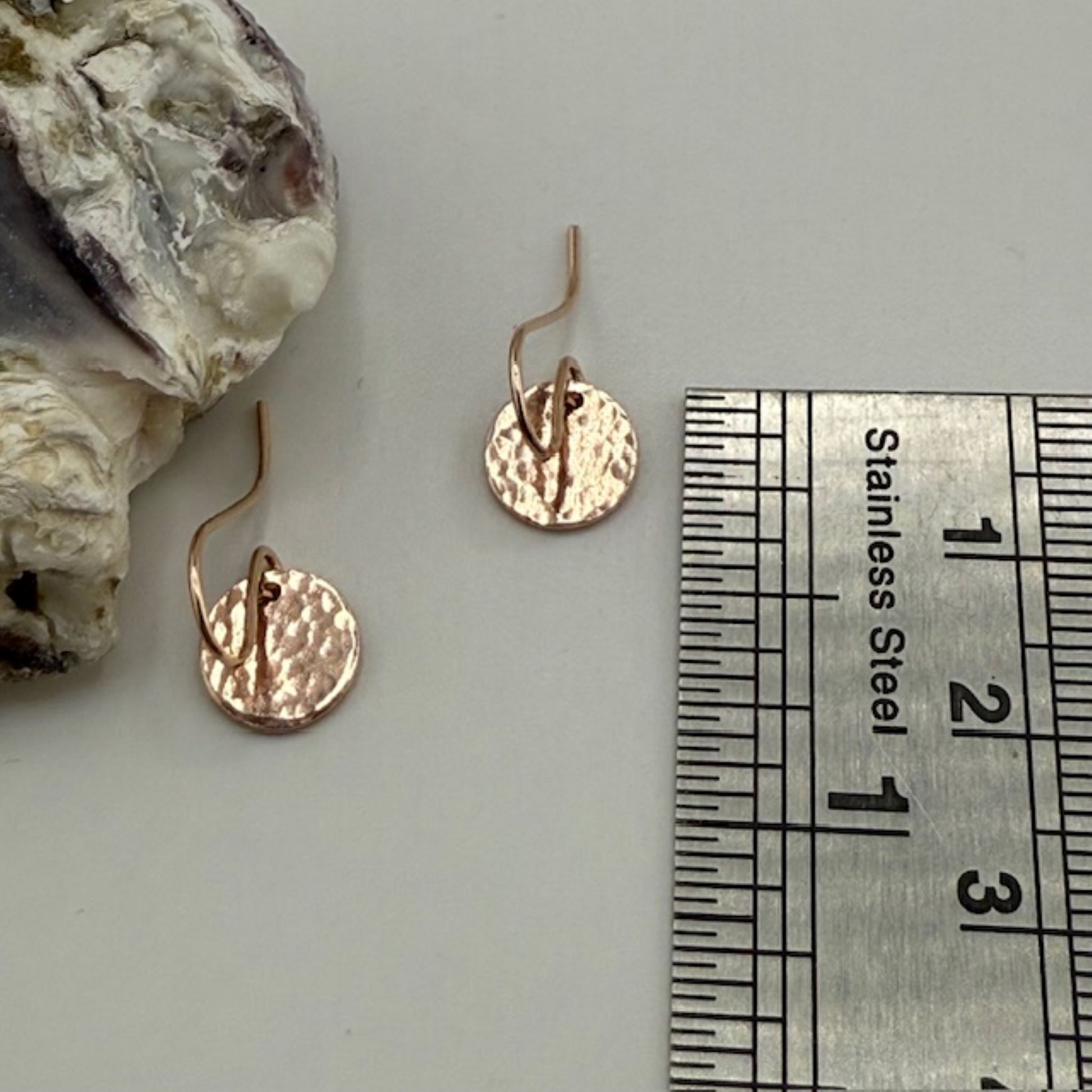 Rose gold earrings next to a ruler for size reference on a neutral background
